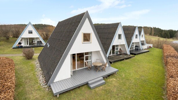 Une rangée de chalets en forme de «A», dotés d’une terrasse et d’une grande fenêtre, situés dans une zone herbeuse, avec des arbres en arrière-plan.