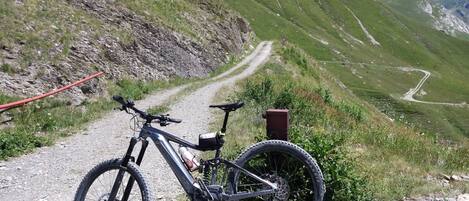 Una bicicleta de montaña aparcada en un camino de grava, con una vista panorámica de una carretera serpenteante y montañas al fondo.