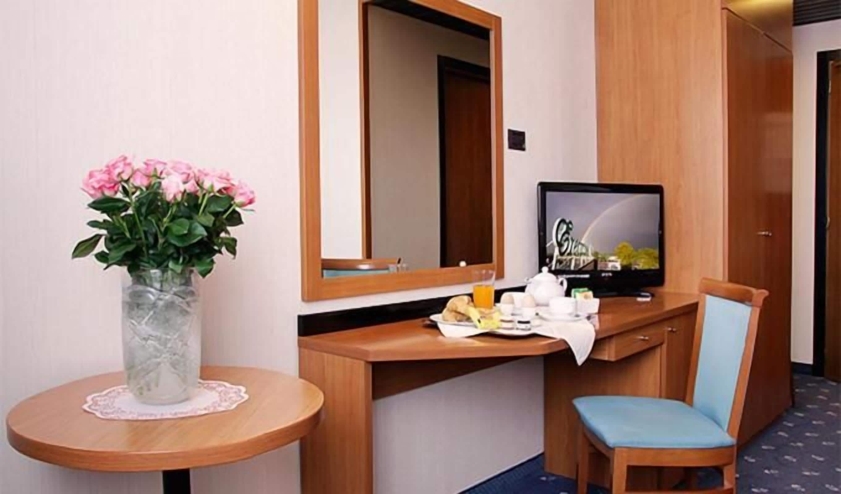 Una camera d'albergo con una scrivania in legno, una televisione, un vaso con fiori e una sedia.