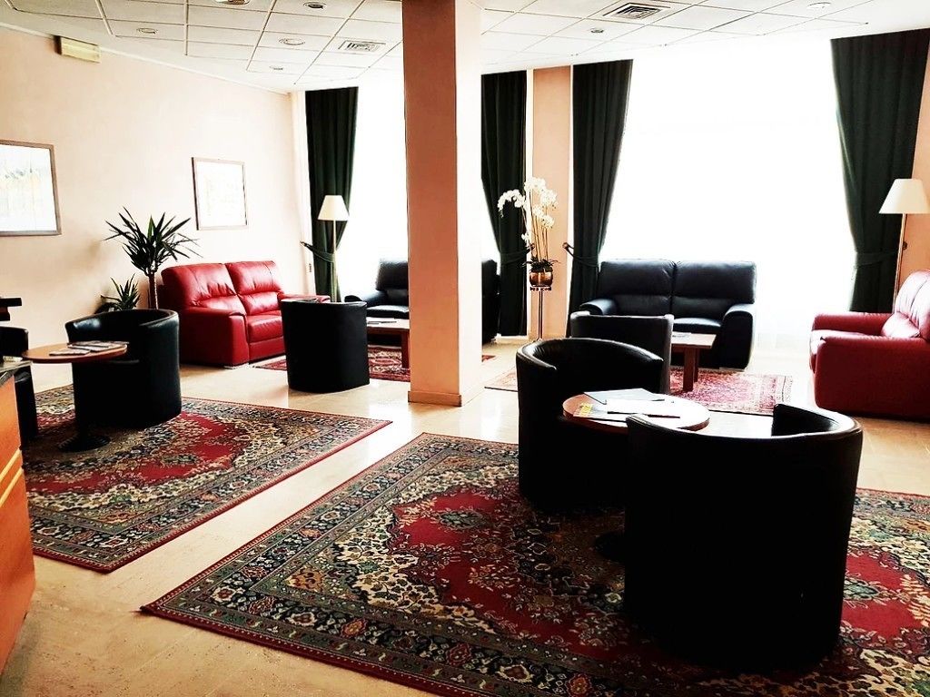 Eine Hotelhalle mit einem roten Ledersofa, schwarzen Ledersesseln, einem schwarzen Ottomanen und einem großen, gemusterten Teppich.