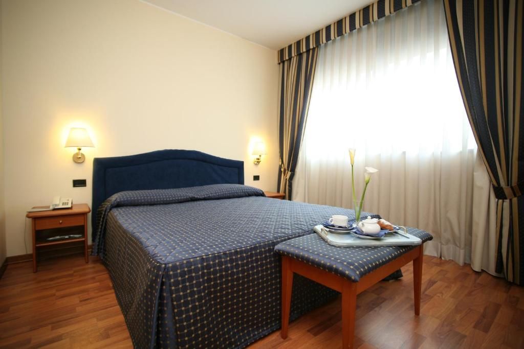 Una camera d'albergo con un letto, abat-jour, un tavolino con vassoio e una finestra con tende.