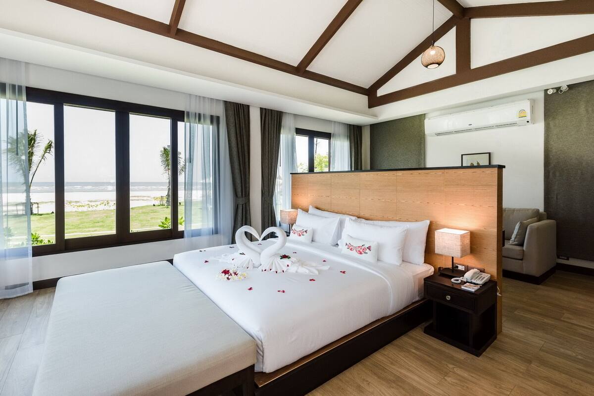 Habitación de hotel con una cama grande, cabecera de madera, mesita de noche con lámpara y vistas a la playa a través de amplias ventanas.