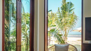 Una habitación con una silla de mimbre, un sofá y un ventanal con vistas a palmeras y una playa.