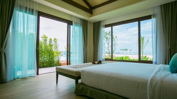 Un dormitorio con una cama grande, un banco y vistas a la playa a través de amplias ventanas.
