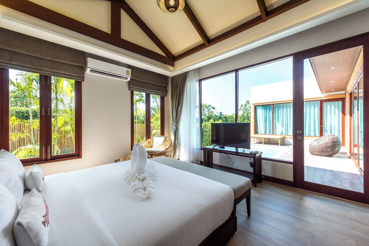 Habitación de hotel con una cama grande, televisión, zona de comedor con mesa y sillas, y ventanas amplias con vistas a zonas verdes.