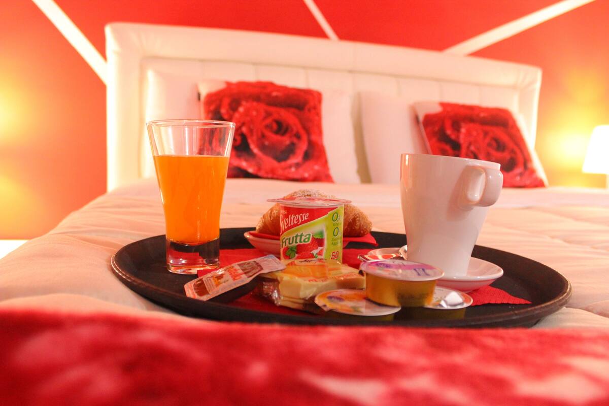 A breakfast tray with a glass of orange juice, a croissant, a cup of coffee, and a container of yogurt on a bed with a white headboard and red rose-patterned pillows.