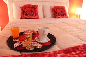 Una bandeja de desayuno ordenada sobre una cama con colcha acolchada blanca, almohadas de rosa roja y una lámpara de noche.