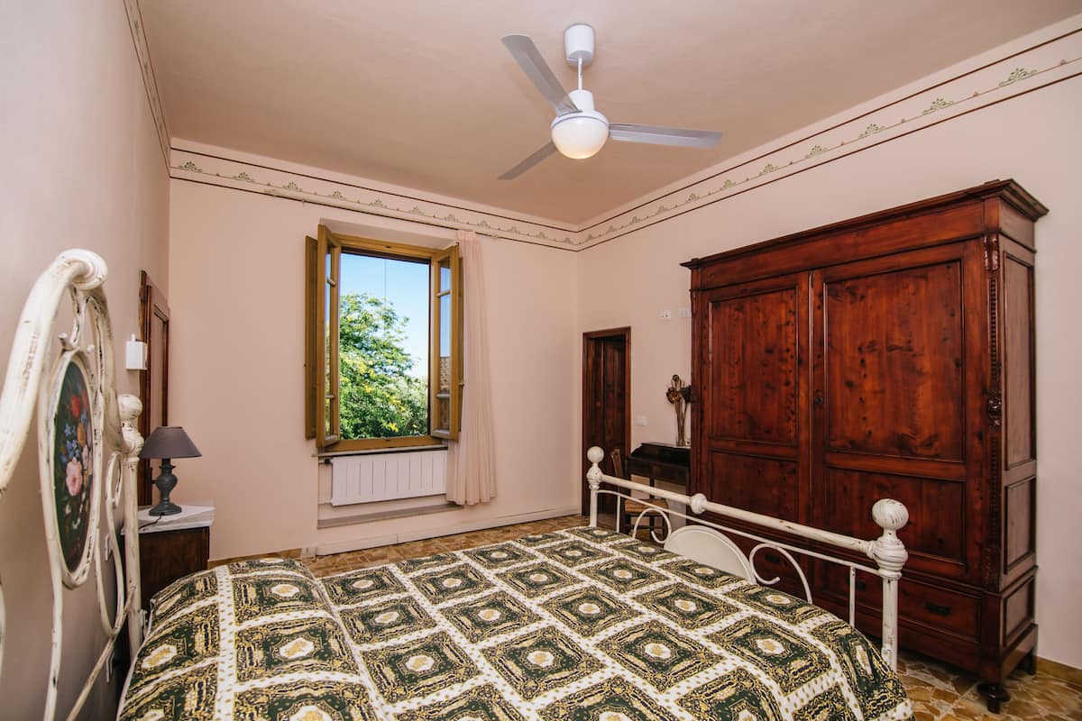 Une chambre à coucher avec une couvre-lit à motifs, une armoire en bois, un ventilateur de plafond et une fenêtre donnant sur un paysage verdoyant.