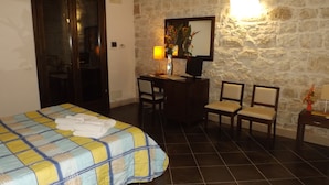 A hotel room with a bed, two chairs, a desk, a lamp, and a stone wall. - Le Baronie (Scicli)
