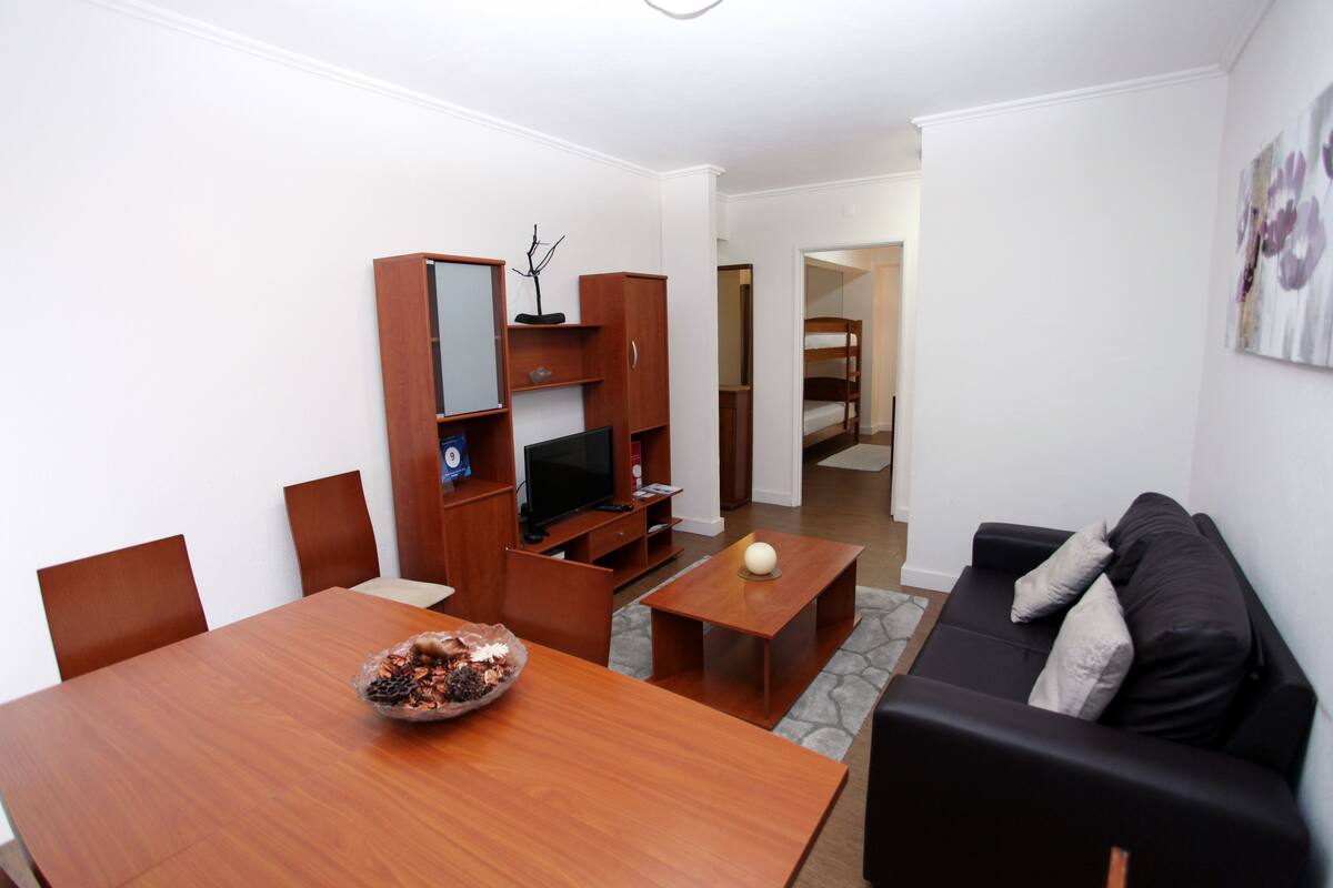 A modern living room with a wooden dining table, a black sofa, and a TV. There is a wooden cabinet with a mirror and a small shelf. The room has white walls and a wooden floor.