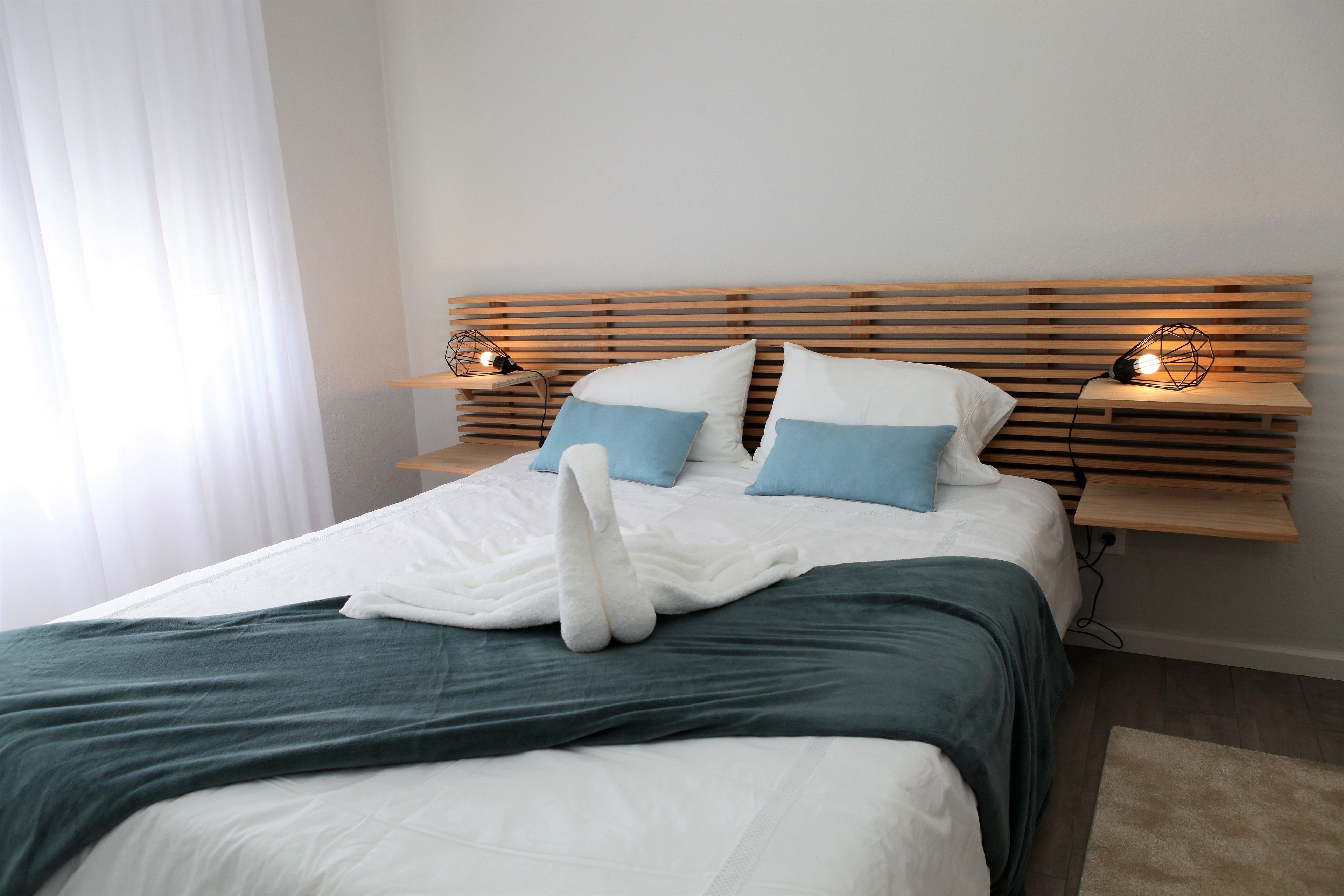 A neatly made bed with a white and teal duvet, two blue pillows, and a white towel on the bed. There are two bedside tables with lamps, and a wooden headboard.