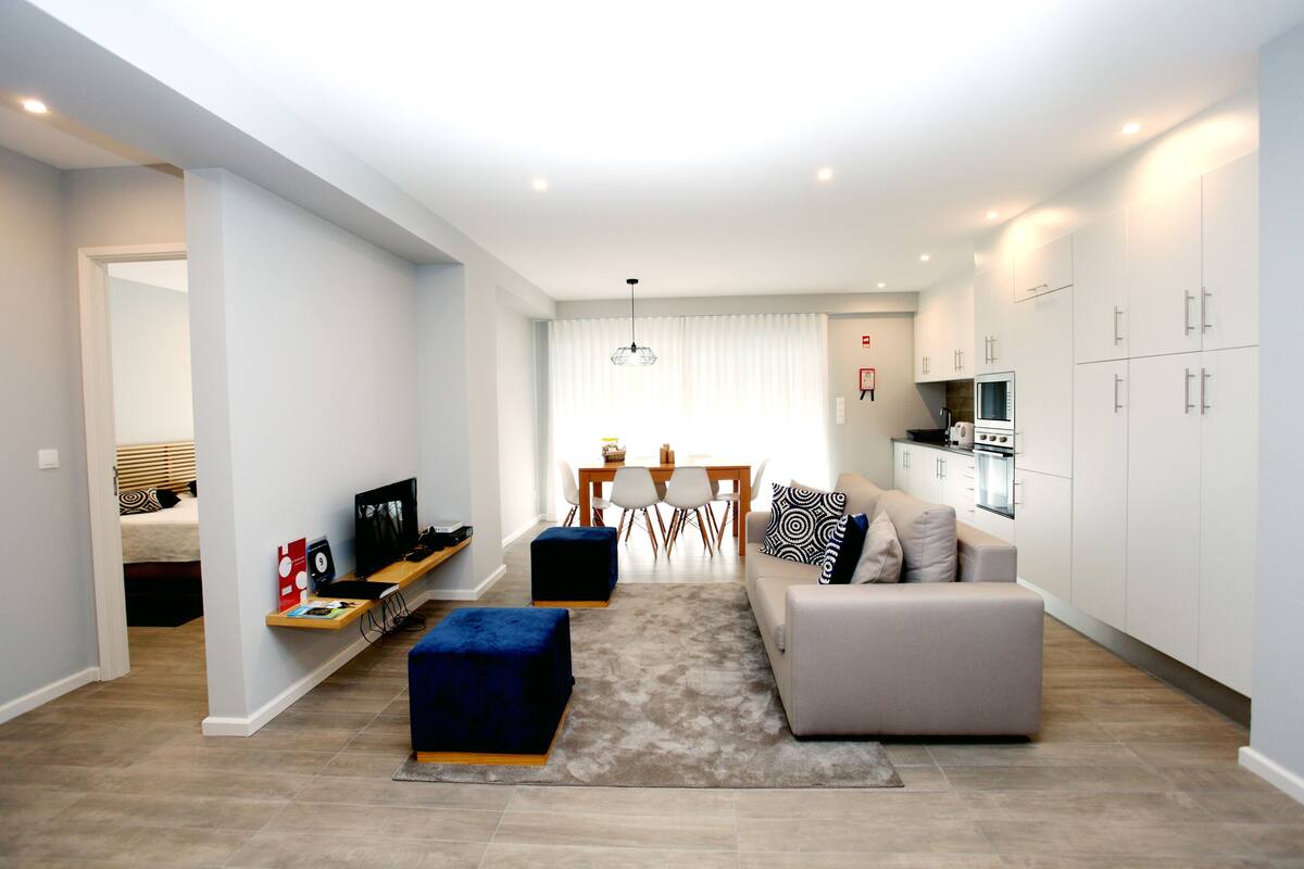 A modern living room with a sofa, ottoman, and a TV on a stand. There is a dining area with a table and chairs in the background.