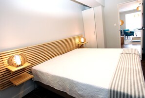 A neatly made bed with a wooden headboard and bedside lamps, a striped bench at the foot of the bed, and a view into a connected room with a blue chair and a lamp. - Apartments 27 Mae de Deus by Green Vacations (Ponta Delgada)
