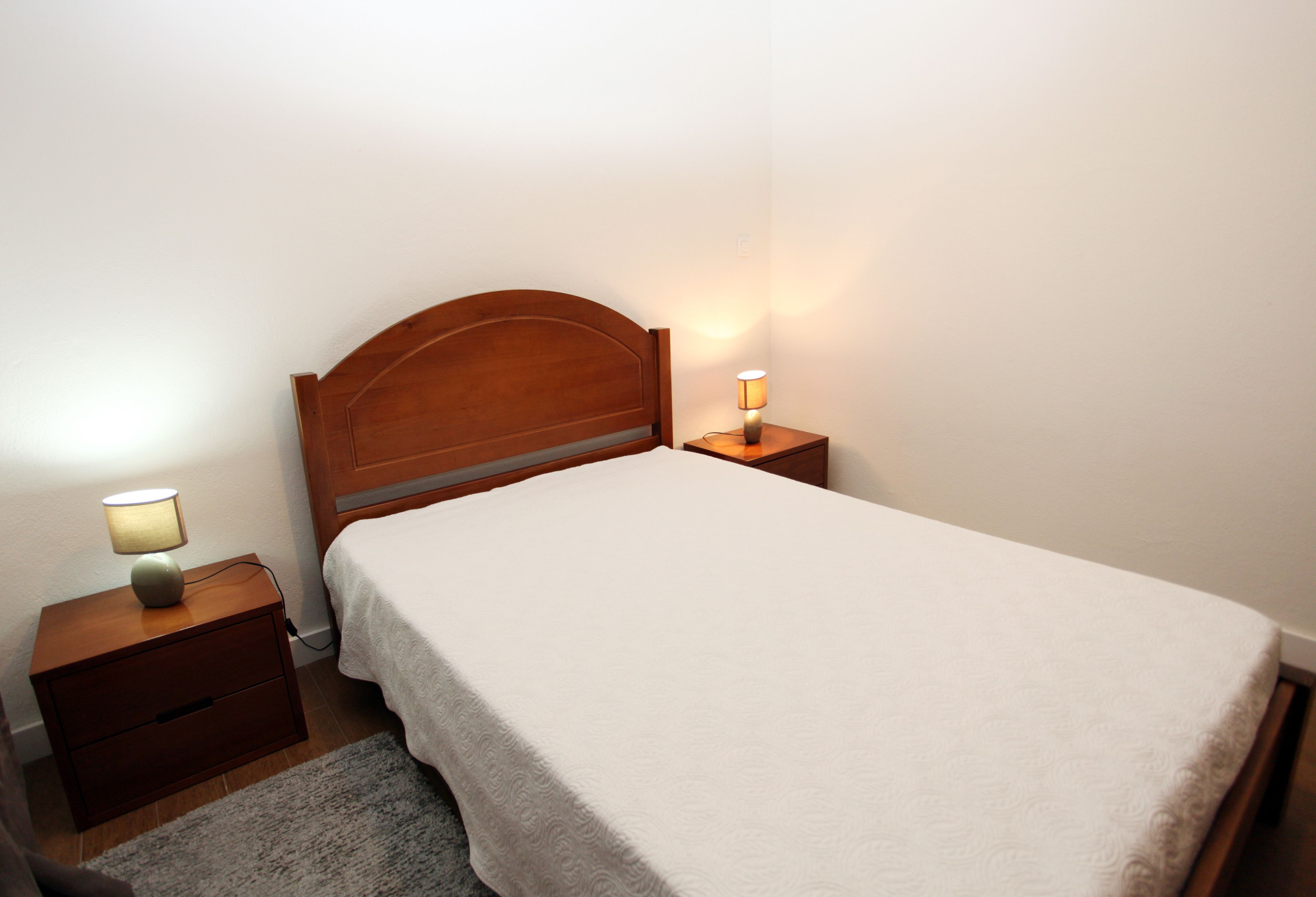A neatly made bed with a wooden headboard, two bedside tables with lamps, and a grey rug on the floor.