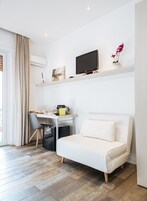 Una habitación de hotel moderna con un sofá blanco, un escritorio con computadora y cafetera, una pequeña área de cocina y un ventanal con cortinas.