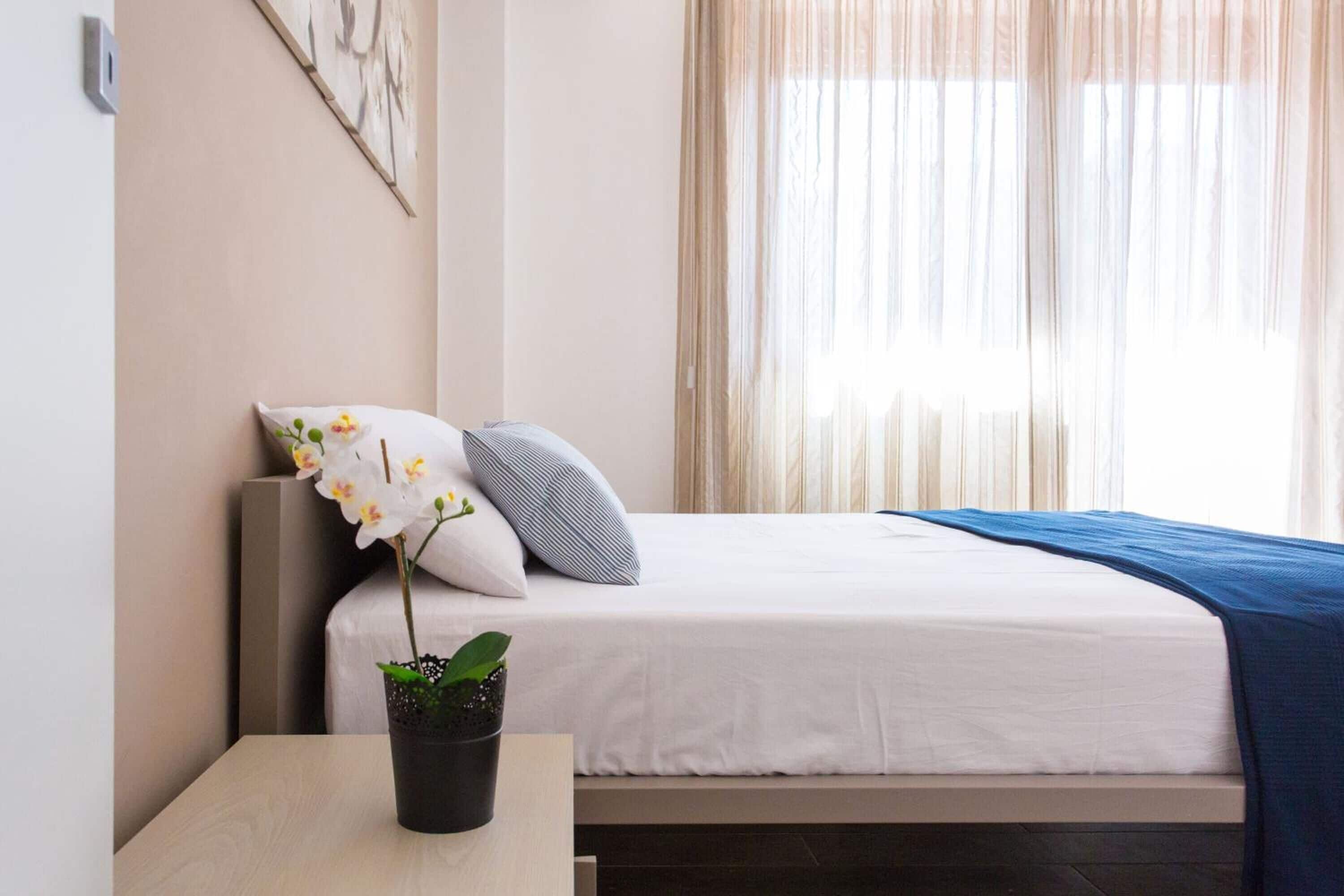 A neatly made bed with white bedding, a blue blanket, and two pillows. There is a small table with a potted orchid beside the bed.