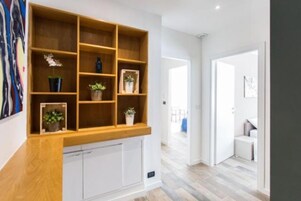 Un interior moderno con una estantería de madera que exhibe plantas en macetas, un mueble blanco en la parte inferior y un pasillo que conduce a otra habitación.