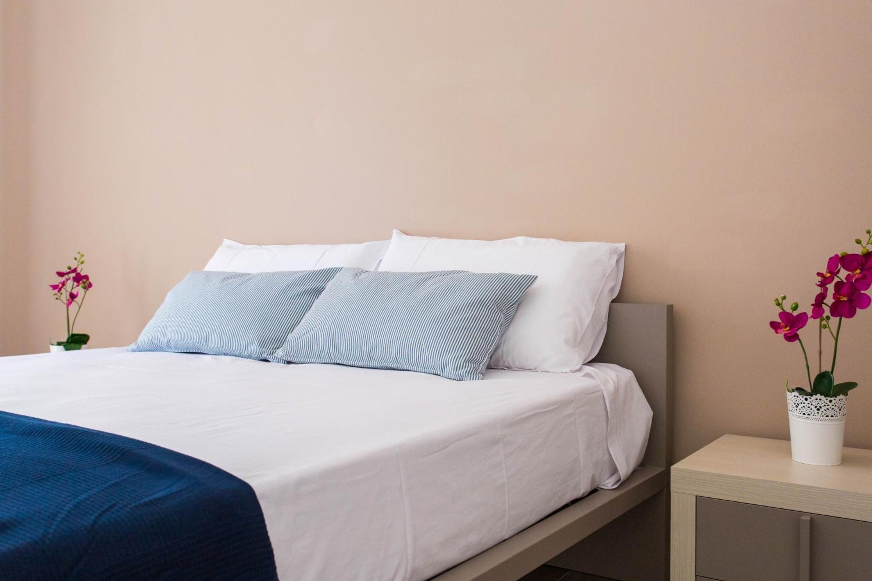 A neatly made bed with white and blue striped pillows, a white duvet, and a blue throw blanket. There is a small bedside table with a white lace pot containing pink flowers.