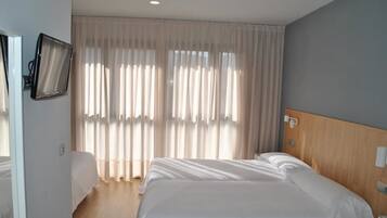 A hotel room with a large bed, a bedside table, a TV mounted on the wall, and a window with sheer curtains.