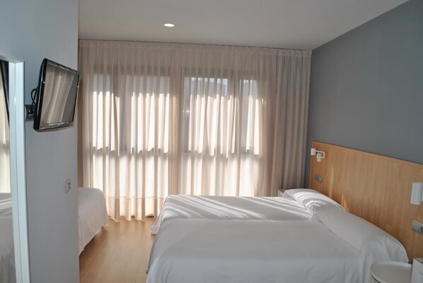 Una habitación de hotel con una cama grande, una mesita de noche, un televisor montado en la pared y un ventanal con cortinas translúcidas.