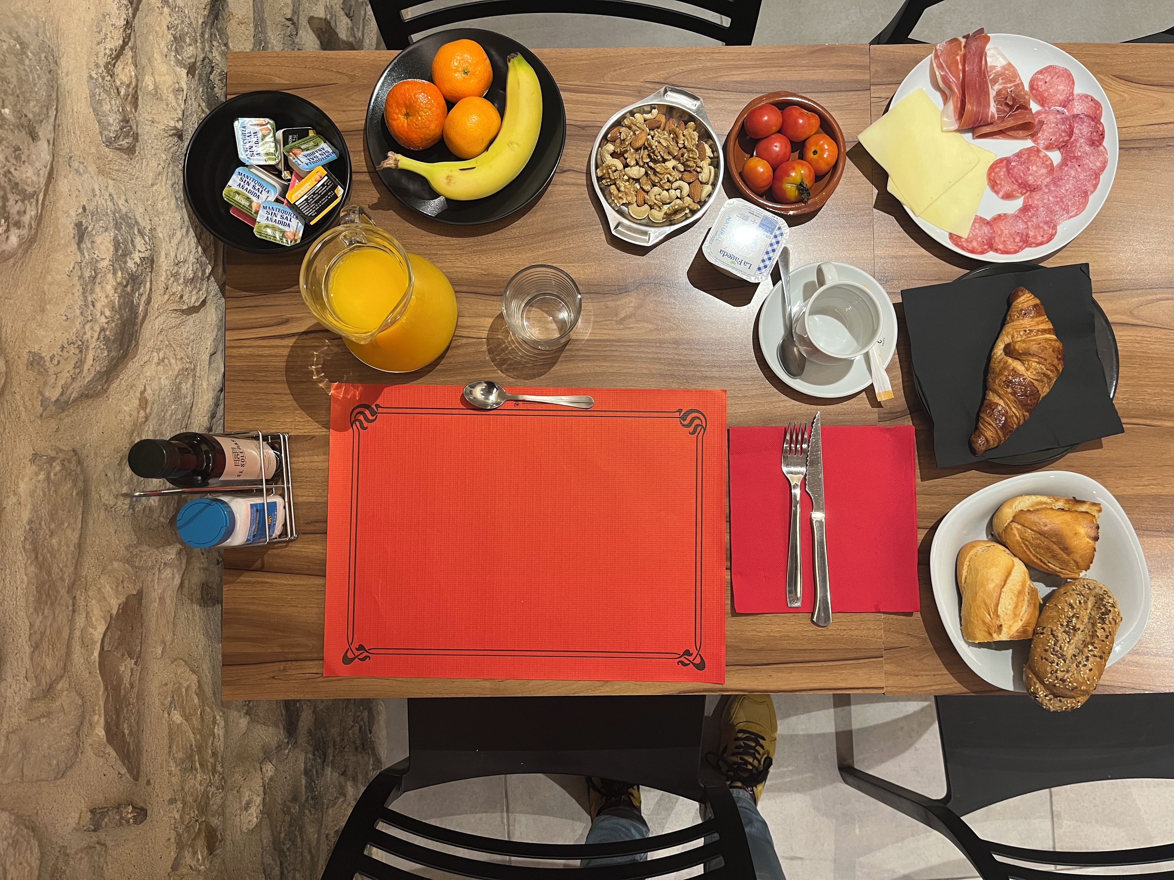 A breakfast table with a variety of food items, including fruits, nuts, cheese, and pastries, along with condiments and a beverage.
