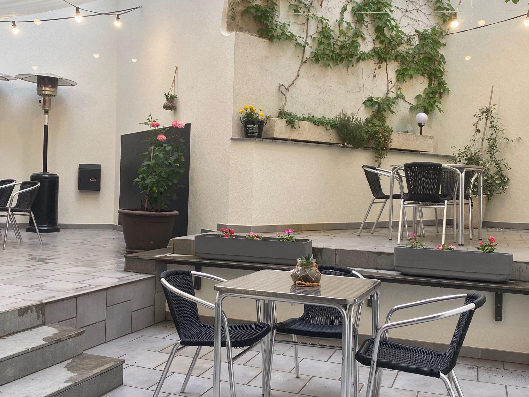 An outdoor patio area with metal chairs, a table, a potted plant, and a wall-mounted shelf with flowers.