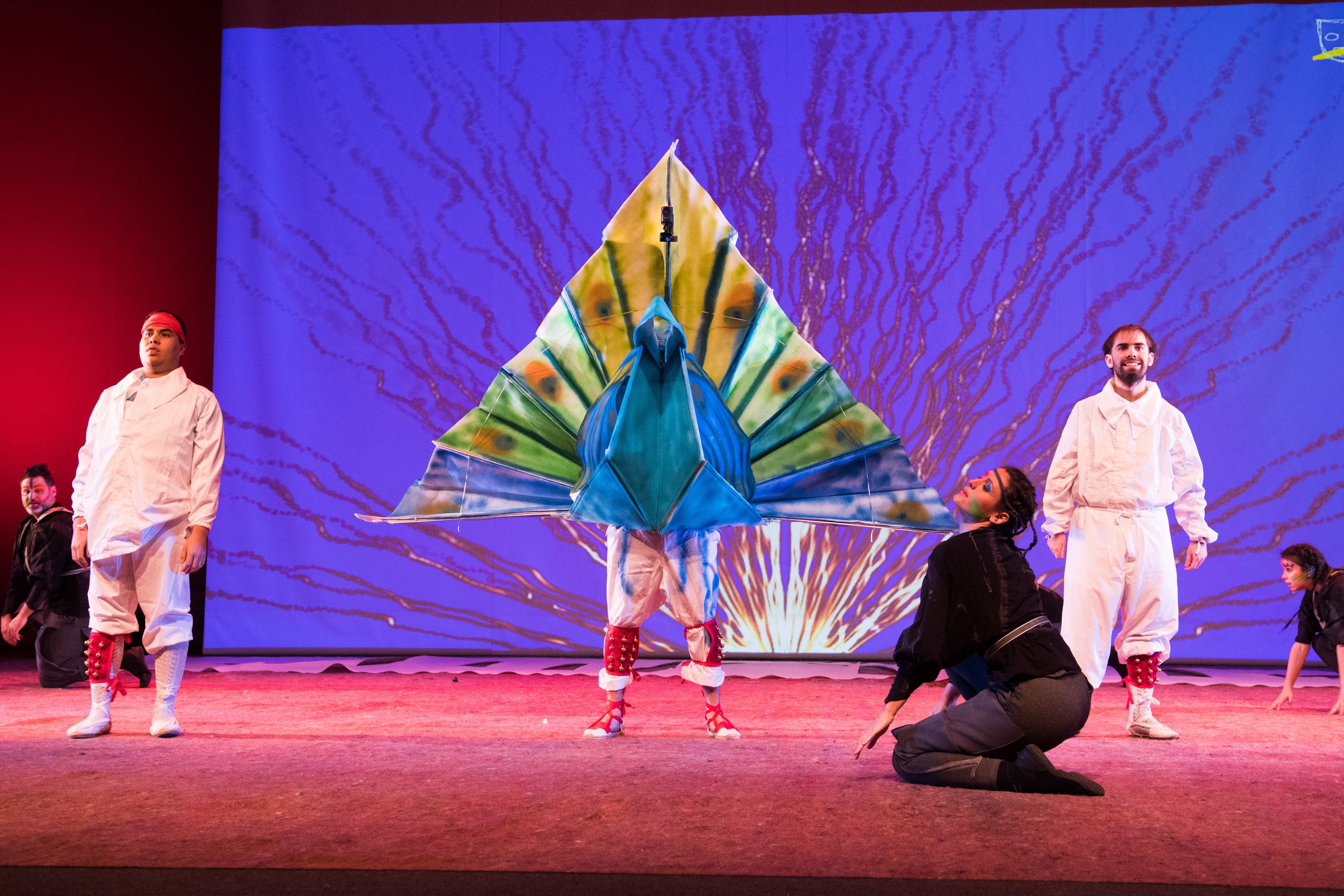 A stage with performers in white costumes, a large peacock feather prop, and a backdrop with a tree-like design.