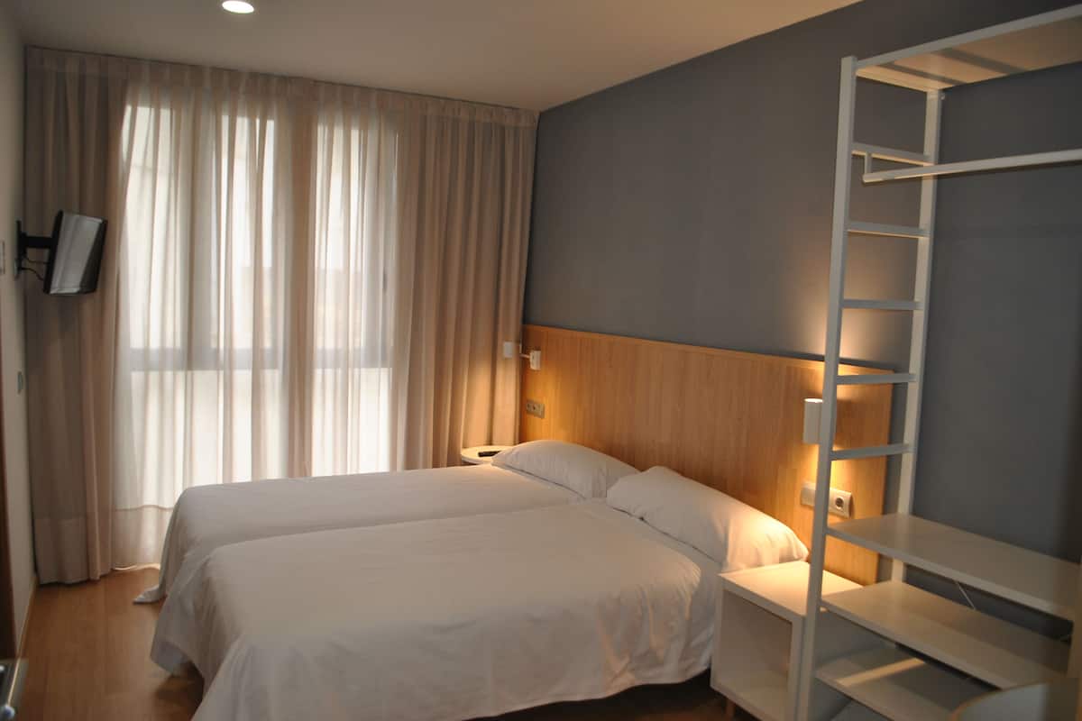 Quarto de hotel com duas camas, cabeceira de madeira, escada branca e uma janela grande com cortinas.