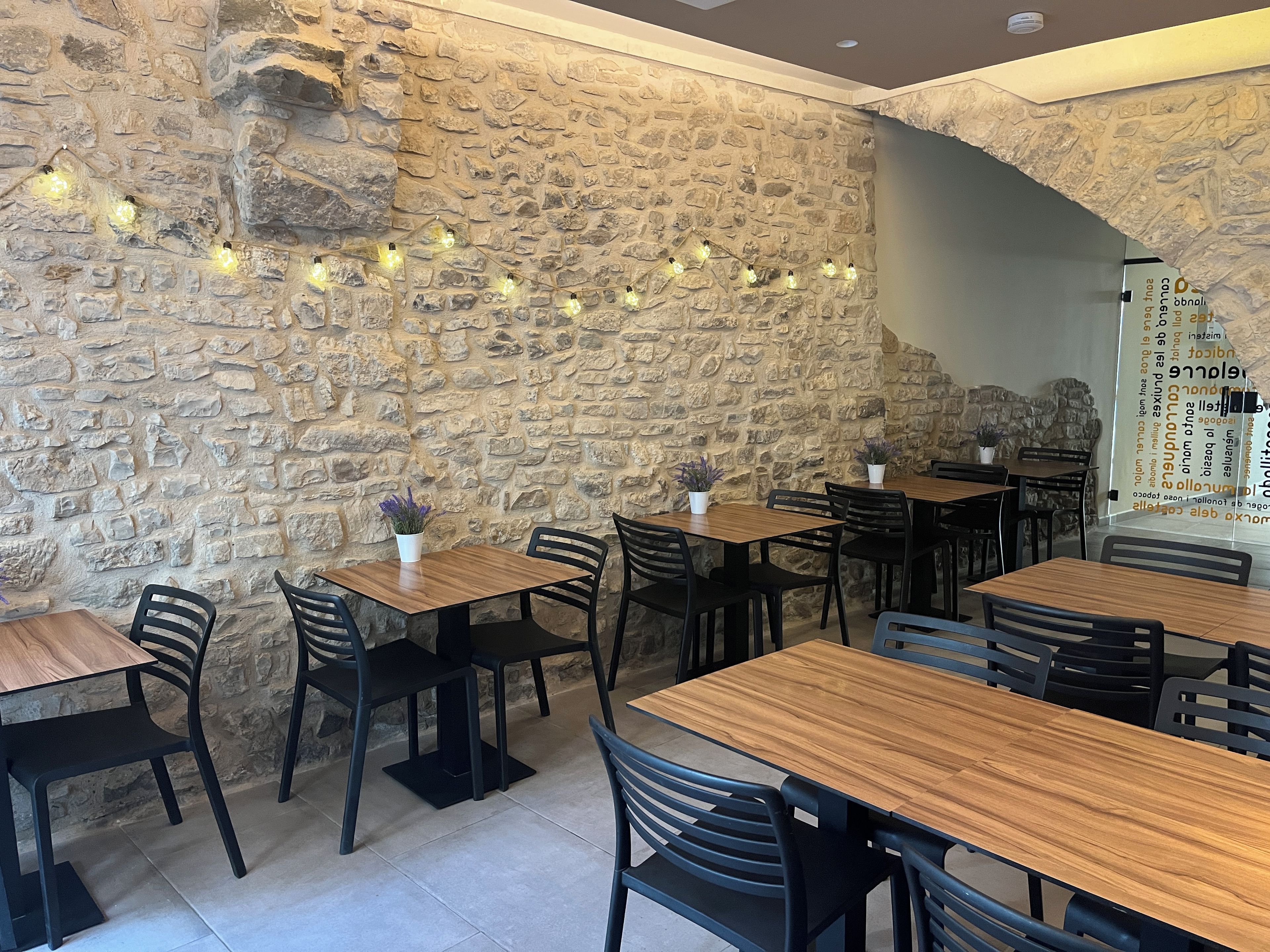 A restaurant interior with stone walls, wooden tables, black chairs, and string lights.