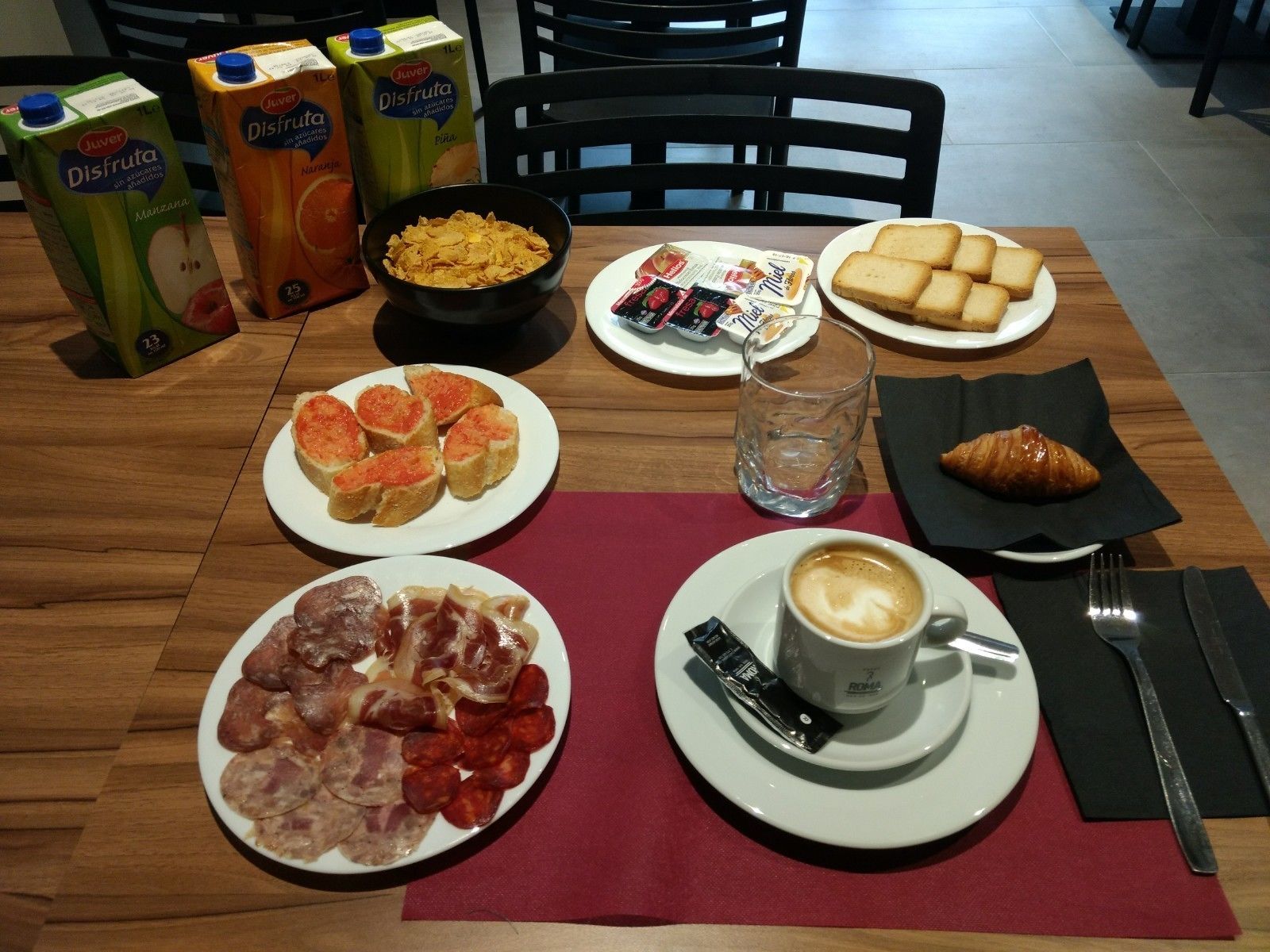 Ett frukostbord med olika sorters mat, inklusive flingor, bröd, bakverk och en kopp kaffe.