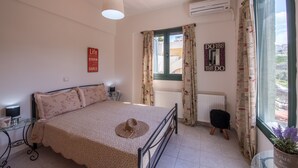 A bedroom with a bed, a bedside table, a lamp, a window with curtains, an air conditioning unit, and a view of the outside. - Ioanna's Home (Malevizi)