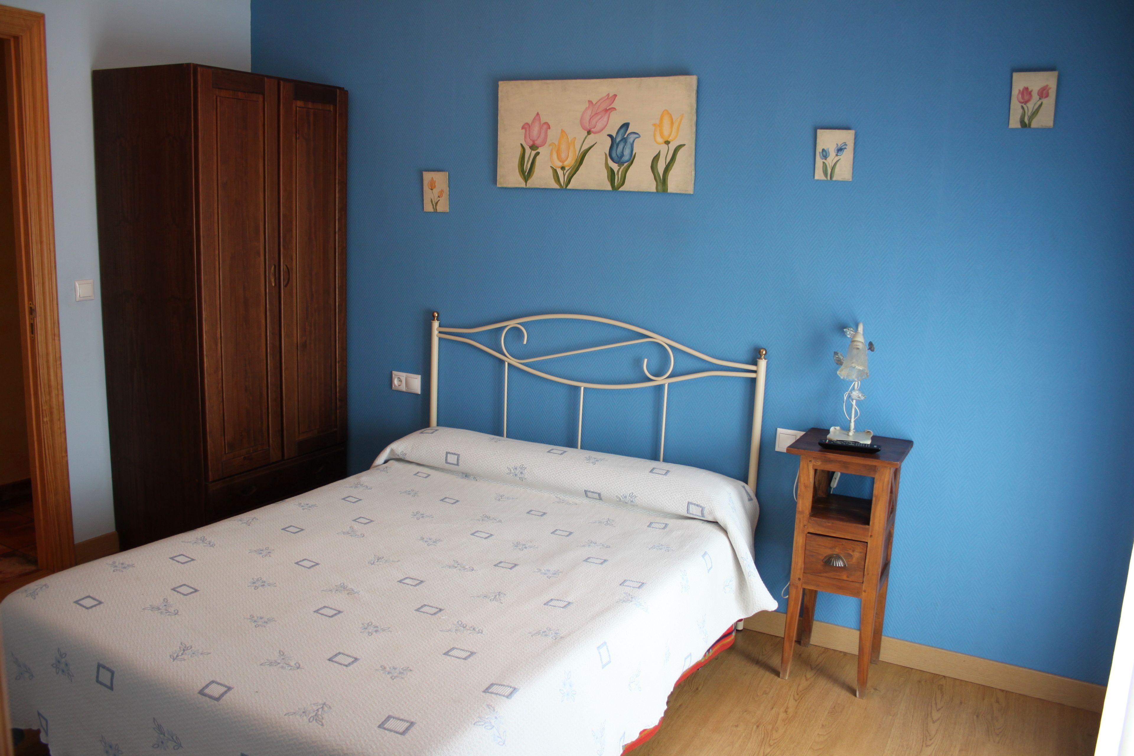 Un dormitorio con cama, armario de madera, mesita de noche con lámpara y un cuadro floral en la pared.