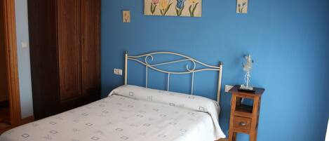 Un dormitorio con cama, armario de madera, mesita de noche con lámpara y un cuadro floral en la pared.