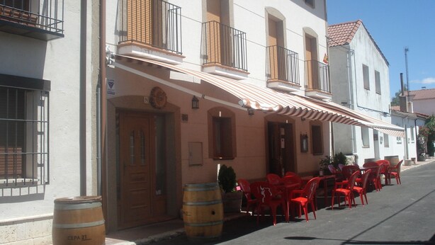 A building with a row of red chairs and tables outside, a wooden barrel, and a door with a sign. - El Rincon del Labrador (Castromonte)