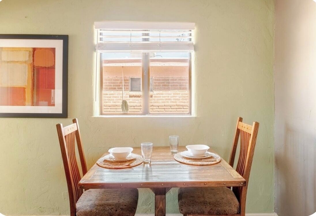 A dining table with four chairs, a window with blinds, and a framed artwork on the wall.