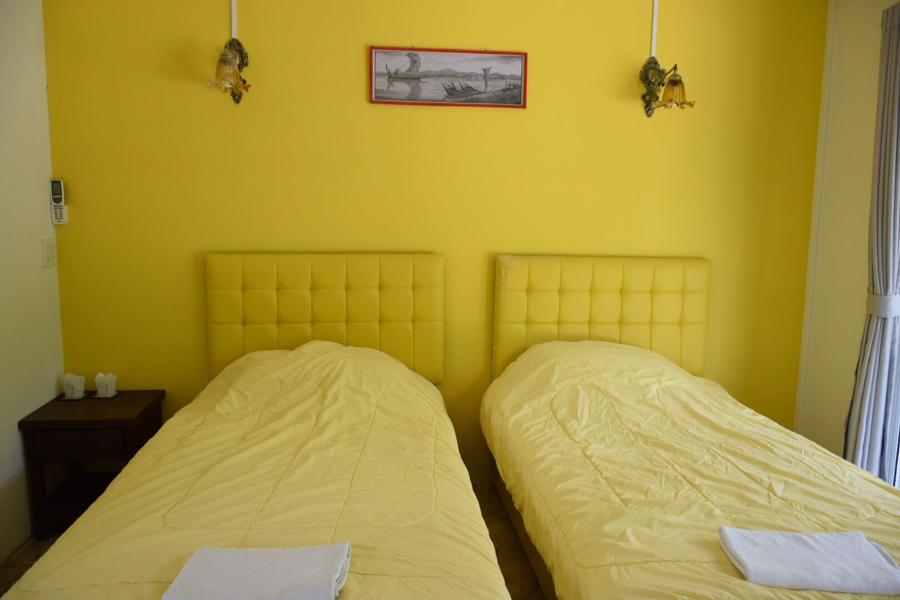 A room with two beds, a bedside table, a wall-mounted lamp, and a framed picture.
