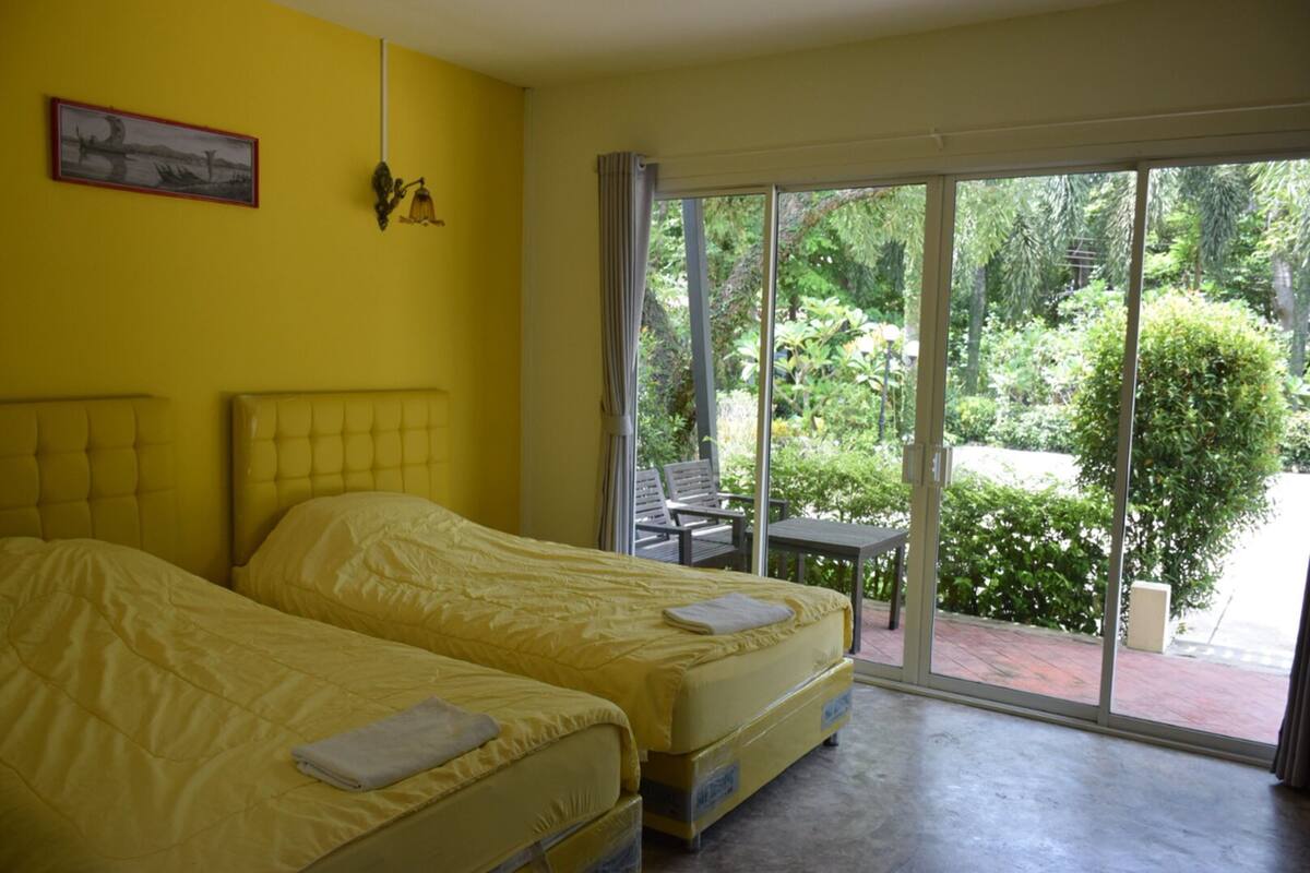A room with two beds, a large sliding glass door leading to a garden, and a small table with chairs outside.