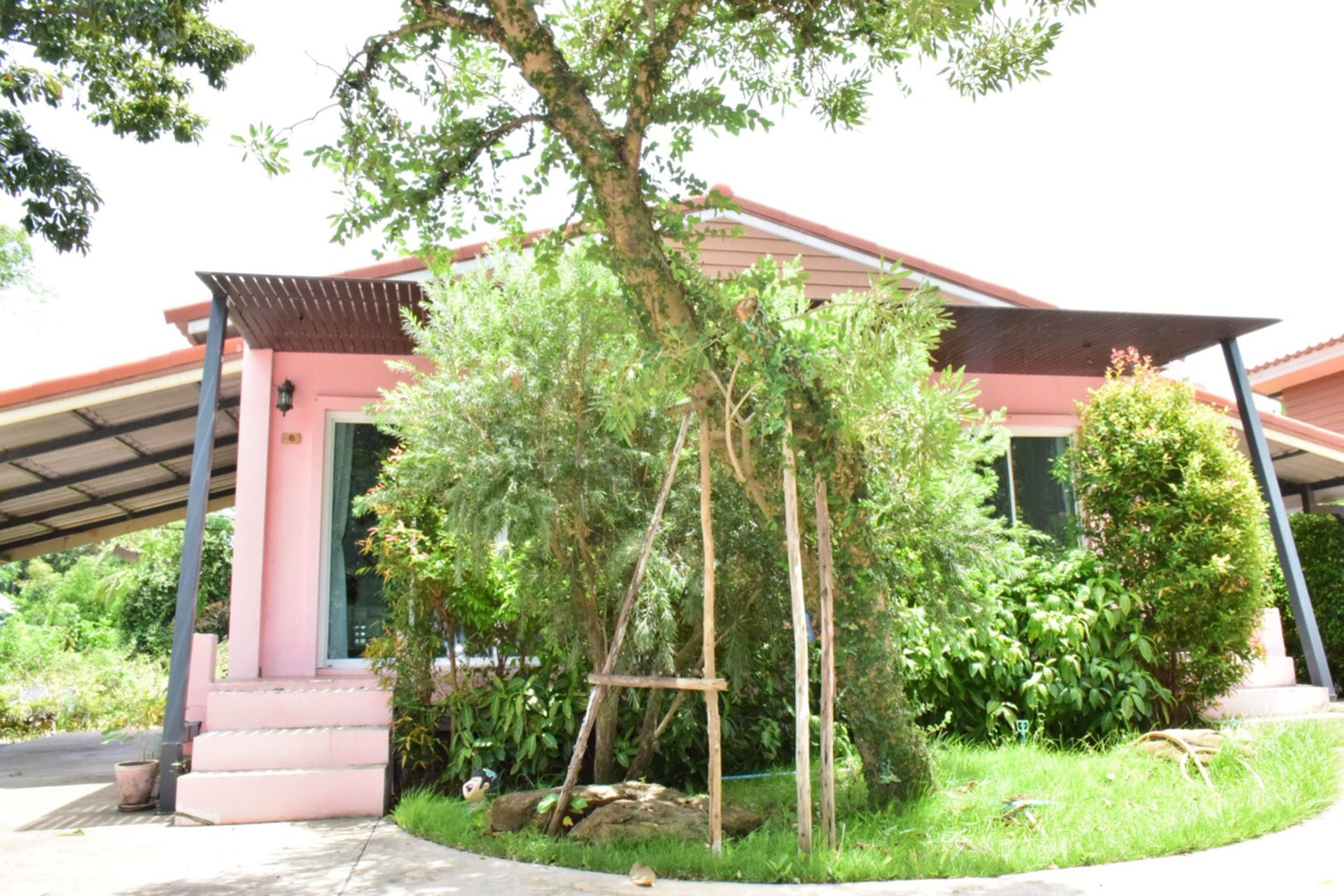 A building with a pink facade, a covered entrance, and a garden area with trees and shrubs.