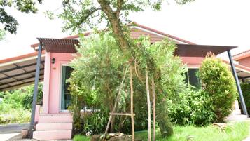 A building with a pink facade, a covered entrance, and a garden area with trees and shrubs.