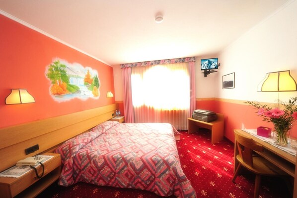 A hotel room with a bed, a desk, a chair, a lamp, a picture on the wall, and a window with curtains. - Hotel Gran Zebrù (Pejo)