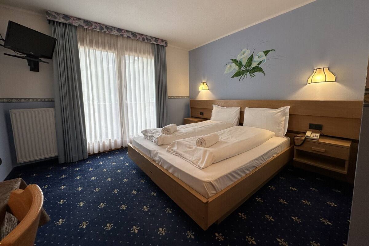A hotel room with a large bed, two bedside tables, a TV mounted on the wall, and a window with curtains.