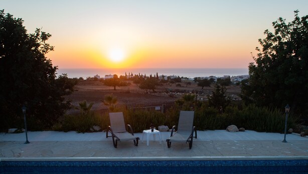 A pool with two lounge chairs facing a sunset view over a landscaped area. - Adonis Villa (Pegeia)