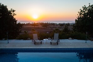 A pool with two lounge chairs facing a sunset view over a landscaped area. - Adonis Villa (Pegeia)