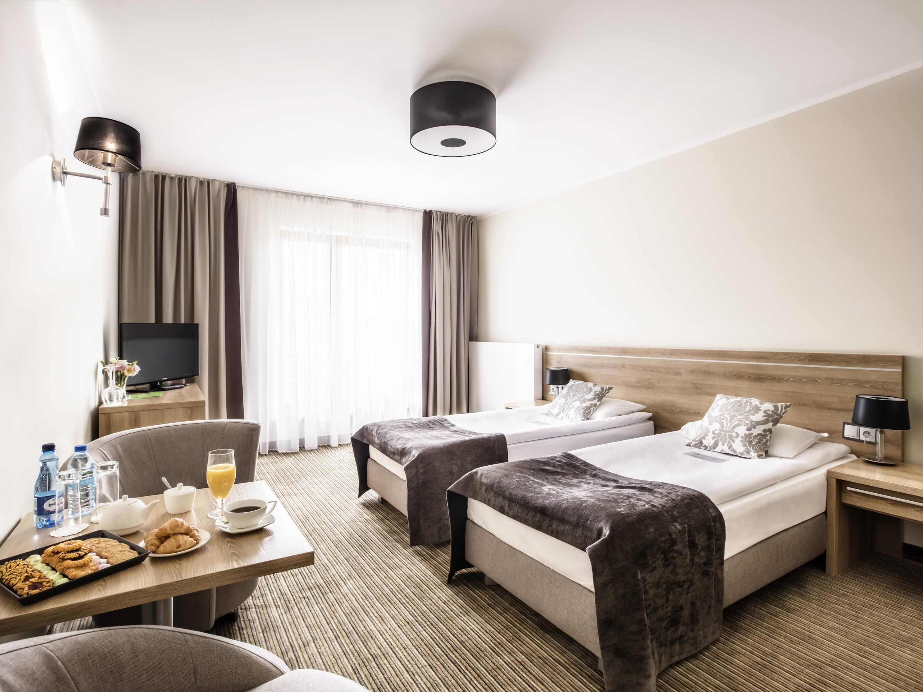 A hotel room with two beds, a TV, a desk with breakfast, and a large window with curtains.