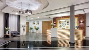A modern reception area with a wooden reception desk, a seating area with a sofa and a floor lamp, and a reception desk with a clock and framed pictures on the wall.