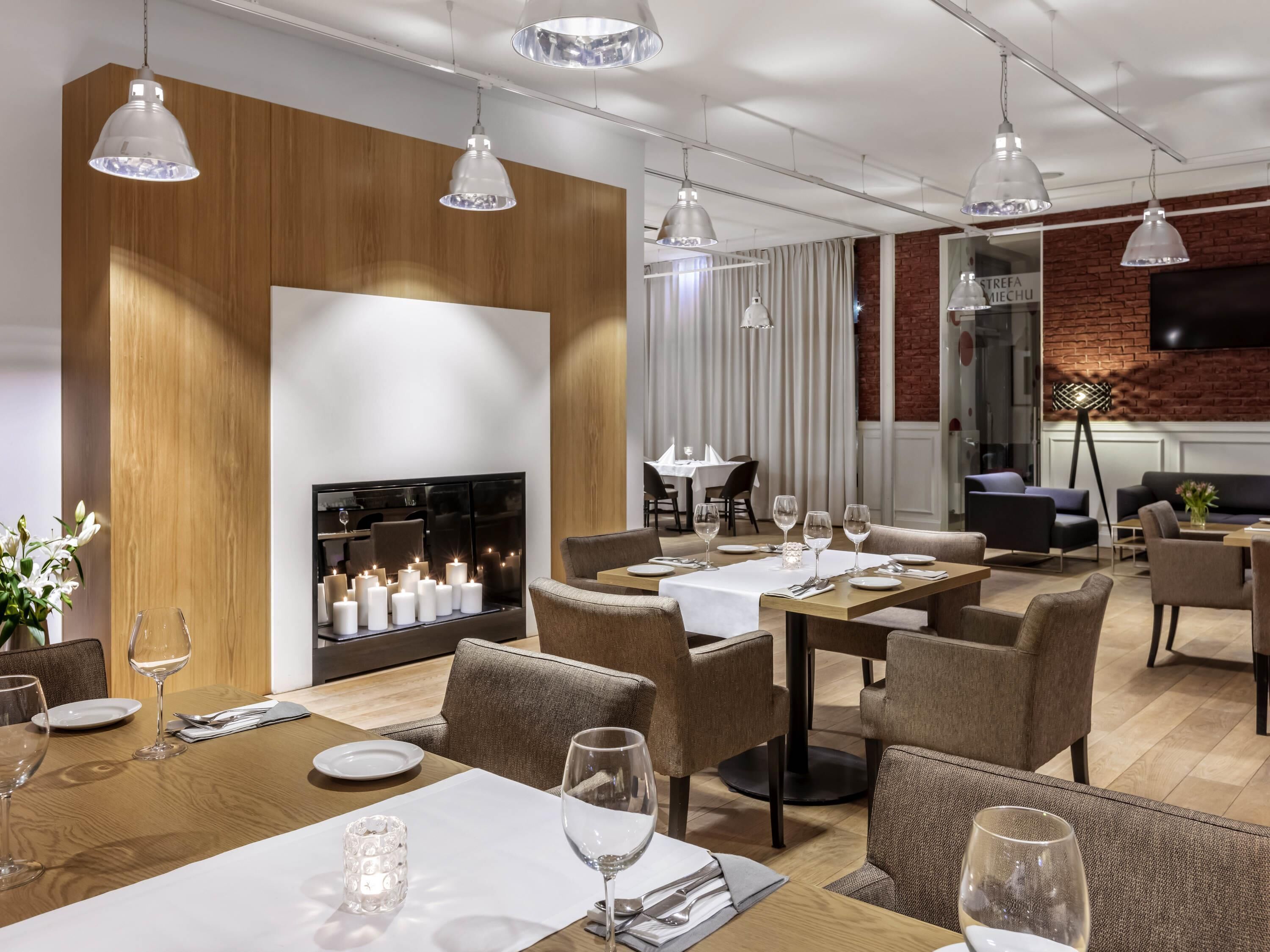A modern dining area with tables set for two, overhead pendant lighting, a fireplace with candles, and a brick wall.