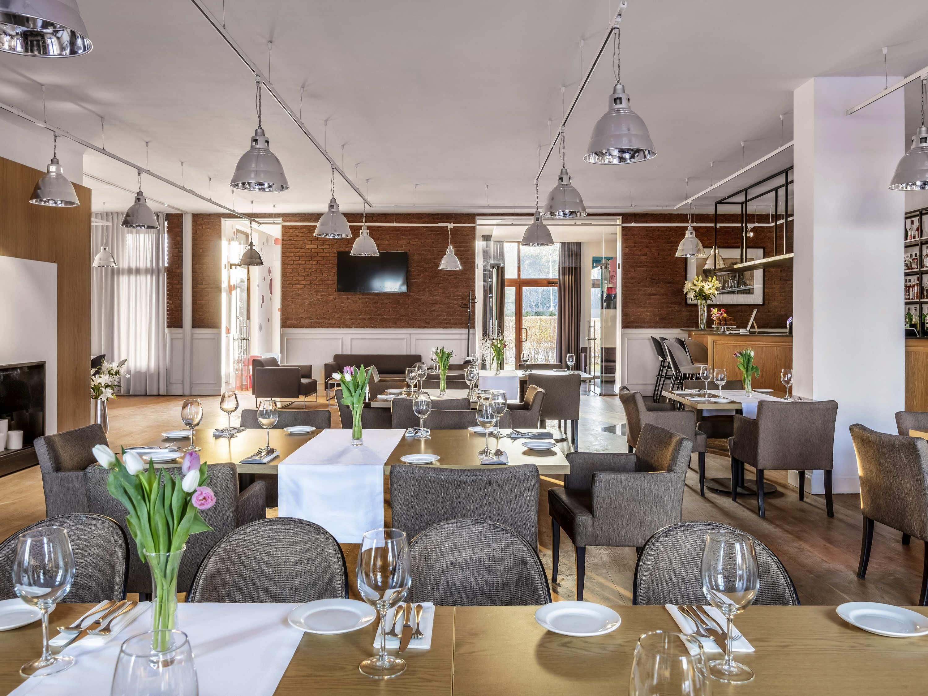 Un restaurante con varias mesas, sillas tapizadas en gris, lámparas colgantes, una pared de ladrillo y un televisor de pantalla plana.