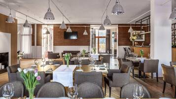 Un restaurante con varias mesas, sillas tapizadas en gris, lámparas colgantes, una pared de ladrillo y un televisor de pantalla plana.