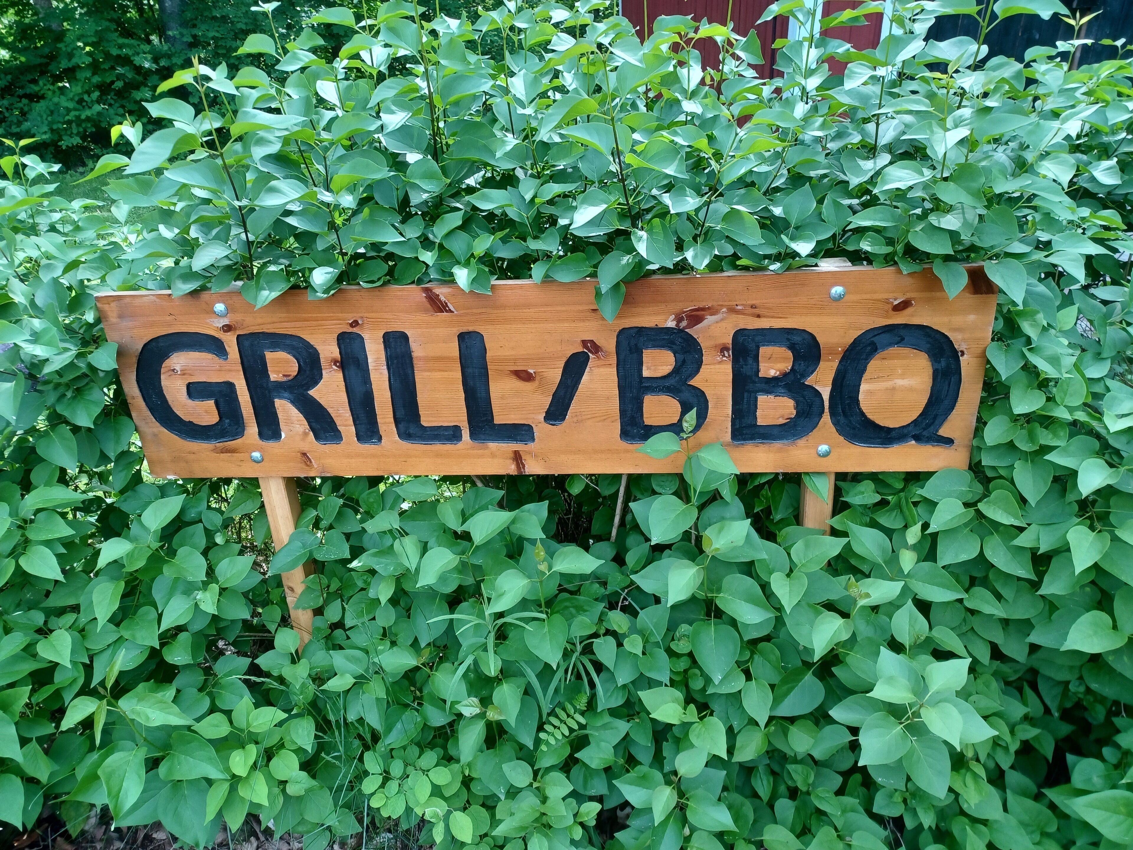 Un letrero de madera con las palabras 'GRILL/BBQ' rodeado de follaje verde y frondoso.