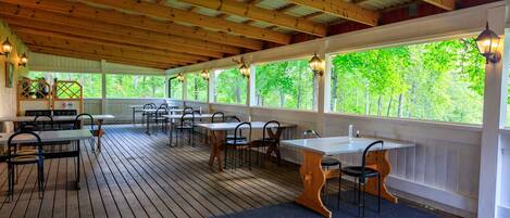 Une terrasse extérieure avec des tables et des chaises en bois, entourée de fenêtres donnant sur la verdure.